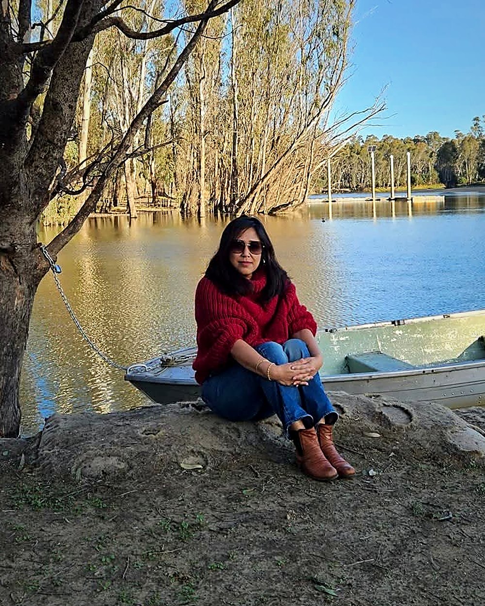 Indira Laisram sitting on a rock by a river, beside a eucalyptus tree and a small boat